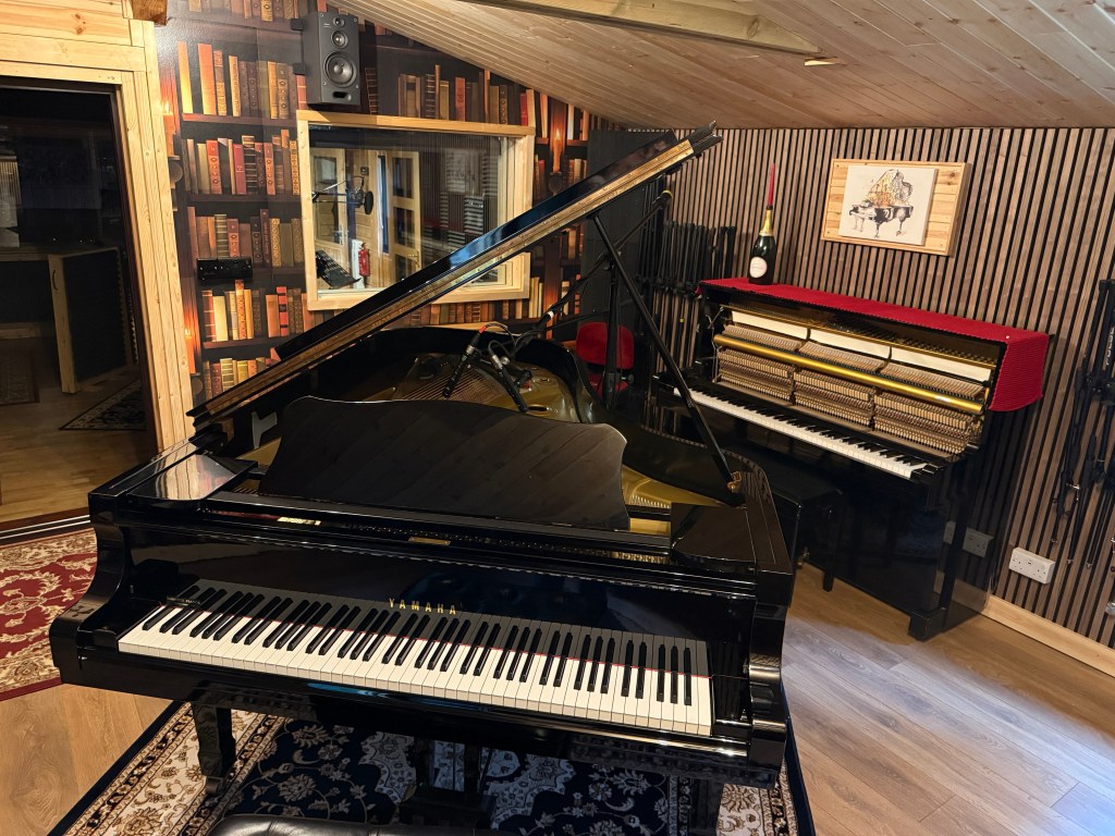 Interior of a music studio featuring a 1979 Yamaha C7 grand piano and a 1968 Yamaha U1 upright piano, with wooden accents and soundproofing elements.