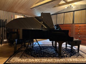 A Yamaha C7 Concert Grand Piano in a recording studio setting, showcasing its polished black finish and positioned beside a smaller upright piano, on a decorative rug under warm lighting.